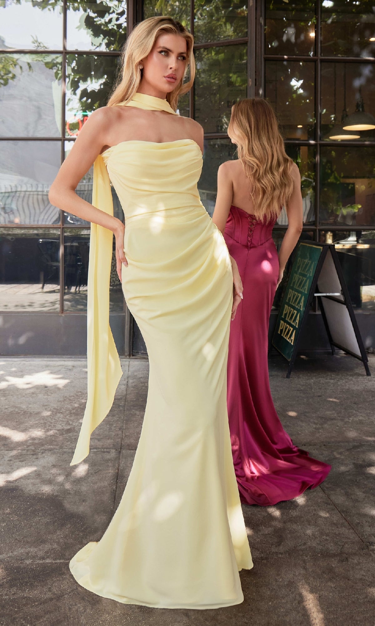 Two women stand outside a restaurant; one wears the Ladivine 7433, a strapless yellow chiffon prom dress with a matching scarf, while the other sports an affordable magenta formal gown. Each is facing in a different direction.